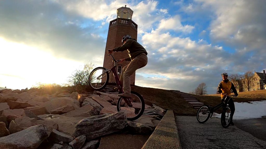 Trials bike rider rear-wheel hopping on rocks with a lighthouse in the background